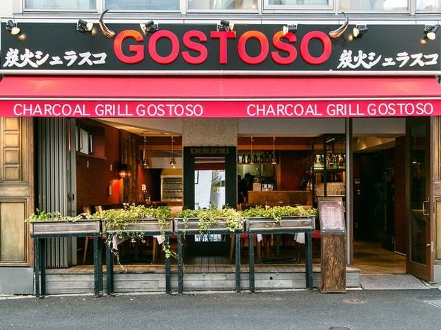 黒い看板に赤い「GOSTOSO」と白い「炭火シュラスコ」、赤いひさしに「CHARCOAL GRILL GOSTOSO」。プランターの緑と開け放したガラスの正面。