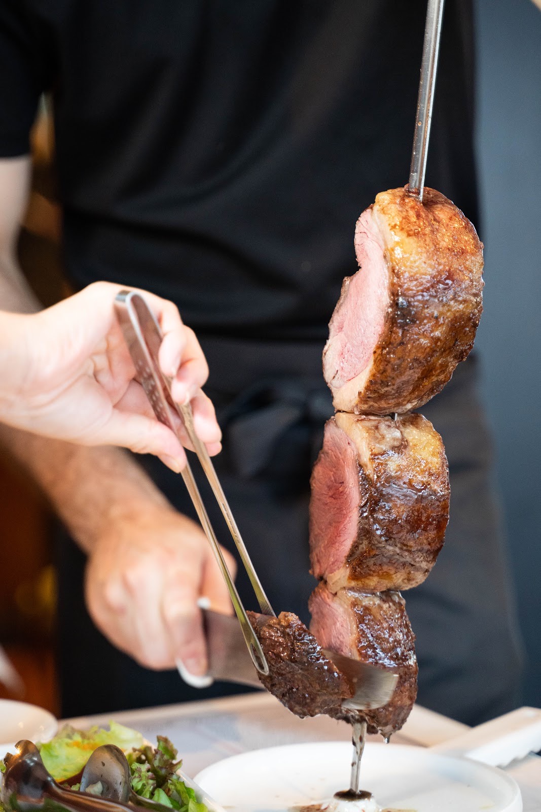 黒いシャツのスタッフが、串に刺した焼き上がりの肉をトングと長いナイフで削ぎ切りにする手元。